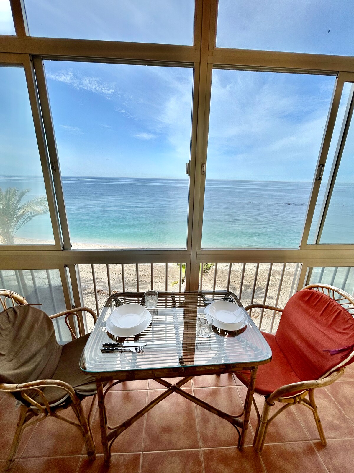 A dining table is situated by large glass windows, providing unobstructed views of the ocean. Two chairs, one in beige and one in red, accompany the table, which is set with two plates. The serene sea and sky are visible beyond the glass.