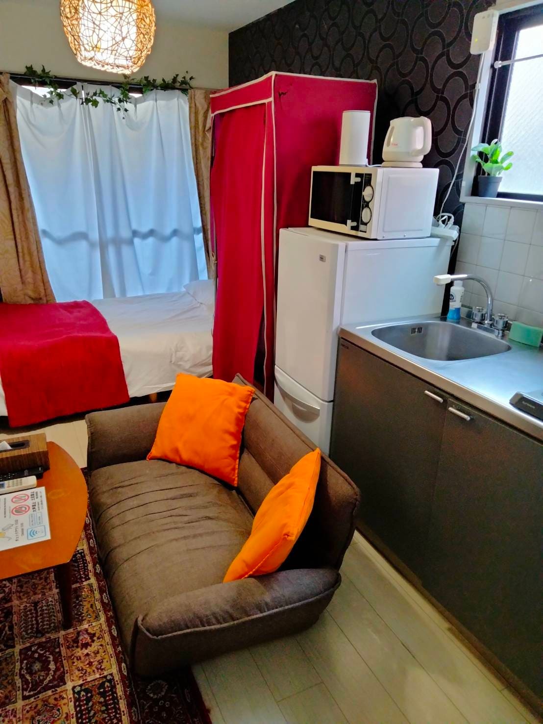 A well-equipped kitchen area is adjacent to a cozy seating space featuring a gray sofa with orange pillows. A compact refrigerator and microwave are positioned on the counter, and a bright bed is partially visible behind a curtain, enhancing the room's functionality.
