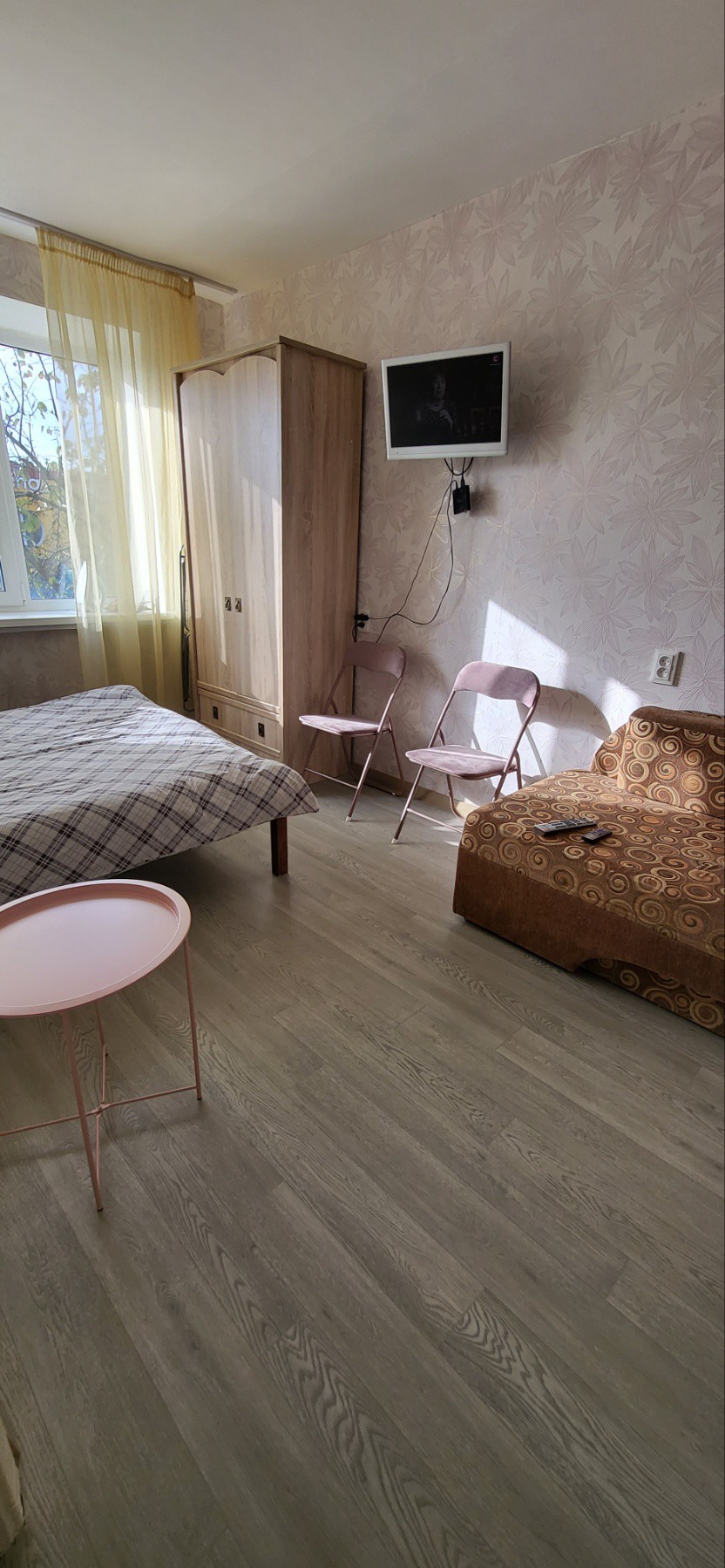 A well-lit bedroom features a spacious bed with a patterned blanket. A small pink side table is positioned beside the bed. Two folding chairs are visible across from a cozy sofa. A wall-mounted television is installed near a window with sheer curtains, allowing natural light to fill the room.