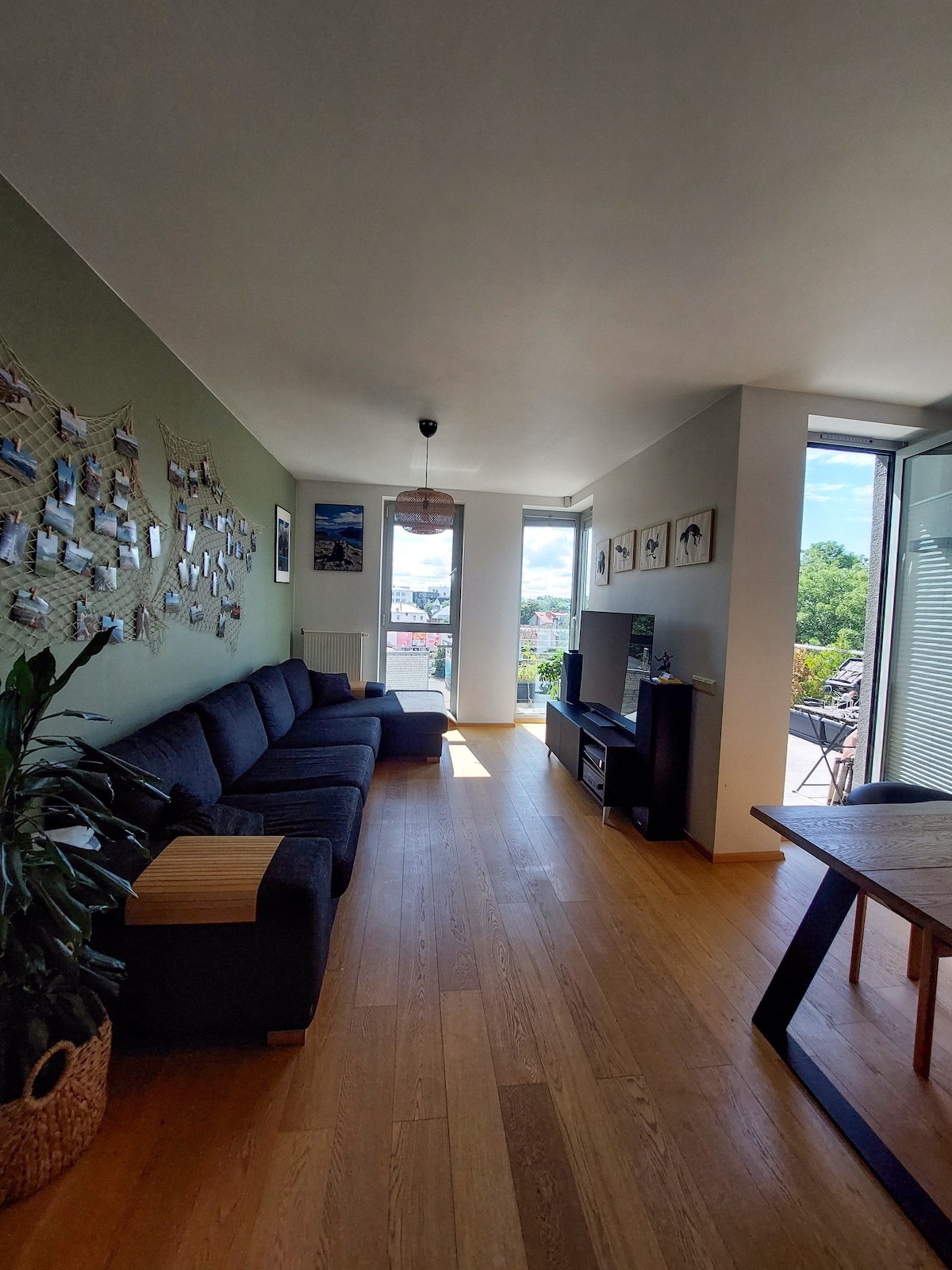 The living area features a spacious couch along one wall, a wooden dining table nearby, and large windows providing natural light and views. A television is positioned opposite the couch, with decorative plants and framed photos enhancing the decor.
