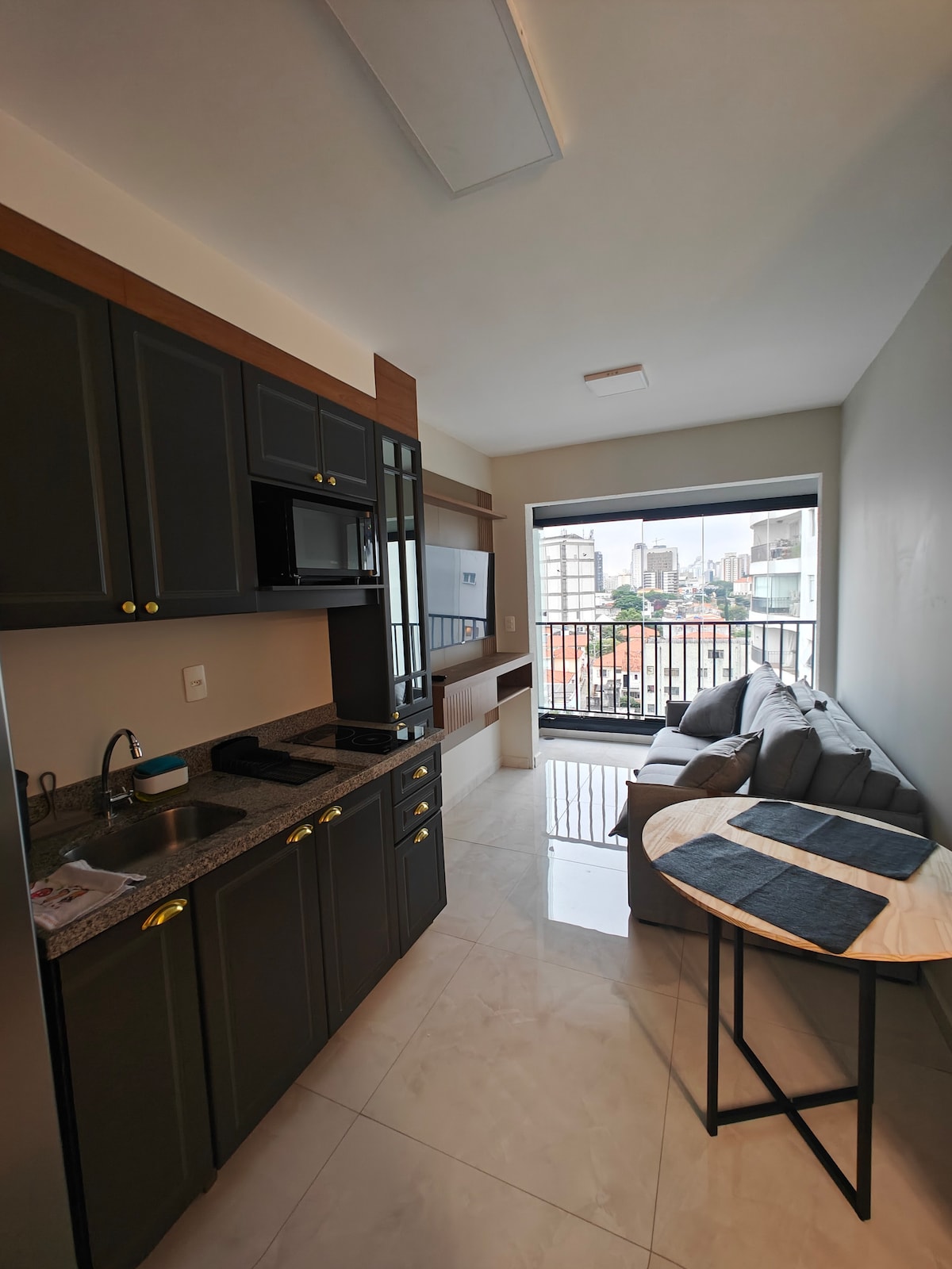 A compact kitchen area features dark cabinetry, a granite countertop, and modern appliances. A cozy seating area includes a light gray sofa and a small round table set with blue placemats. Large windows provide natural light and a view of the city.