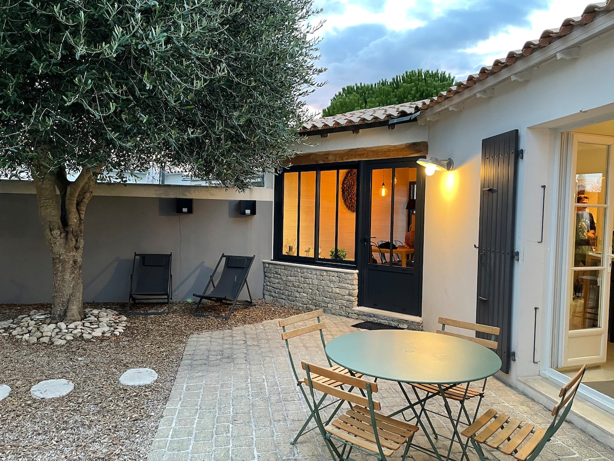 A cozy outdoor seating area features a round green table surrounded by wooden chairs, set on a stone pathway. A decorative olive tree provides shade, while large windows of the adjoining room invite natural light. The space combines tranquility with a welcoming atmosphere.