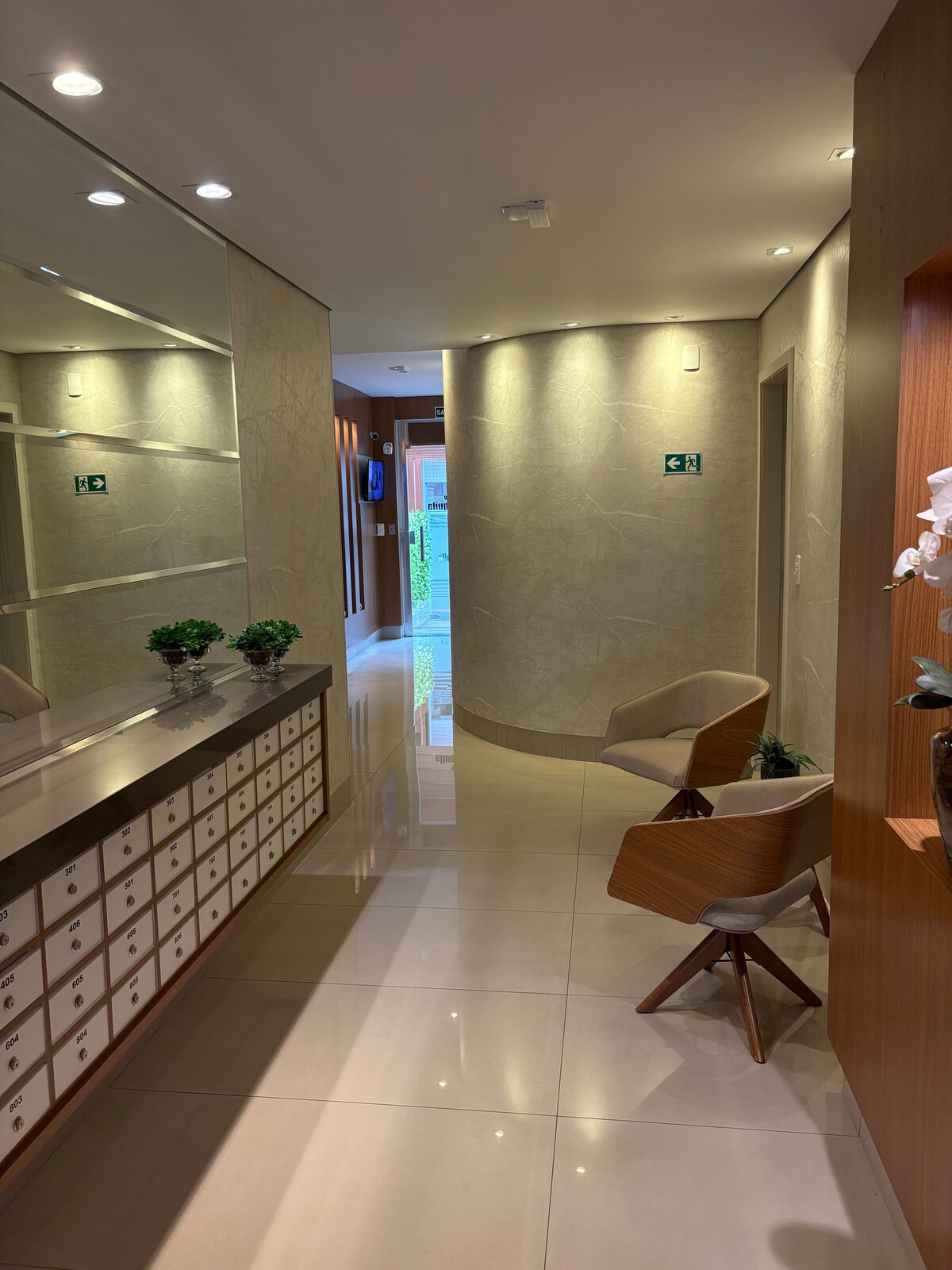A well-lit corridor showcases elegant marble walls and a mirrored vanity. Stylish wooden chairs are positioned beside a decorative table with small potted plants. The smooth, tiled floor reflects the warm lighting, leading to a glimpse of outdoor greenery through a glass door at the end.