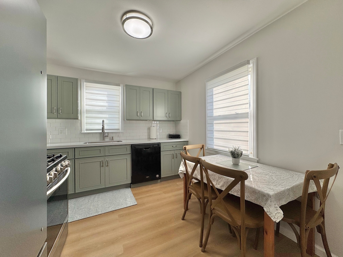 A modern kitchen is presented, featuring gray cabinetry and a spacious countertop. Natural light enters through two windows, illuminating a cozy dining area with a wooden table and four chairs. Stainless steel appliances, including a gas stove, are visible, enhancing the kitchen's functionality.