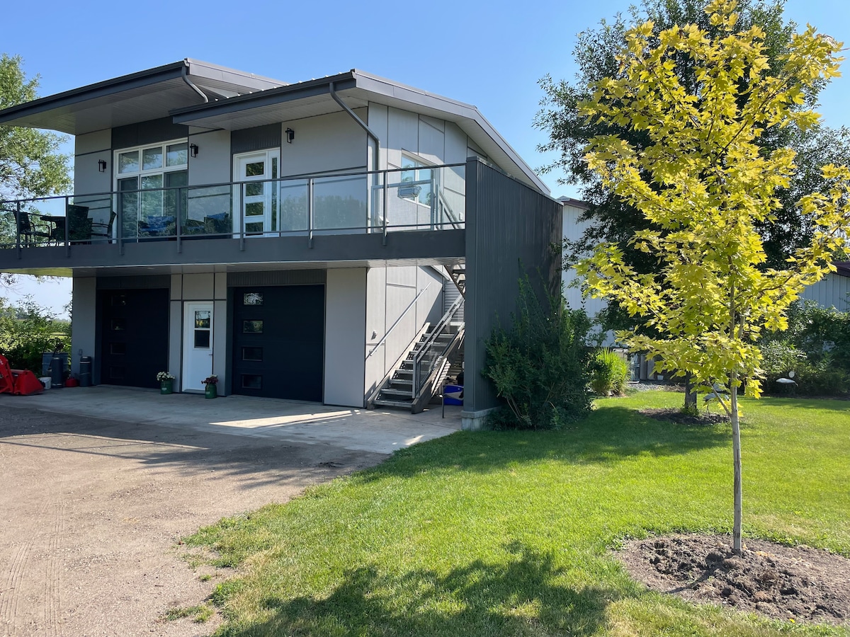 A modern loft is perched above a two-car garage, featuring a spacious balcony with glass railing. The exterior is complemented by a neatly maintained lawn and a single small tree. Clear skies provide a bright backdrop, highlighting the architectural simplicity of the building.