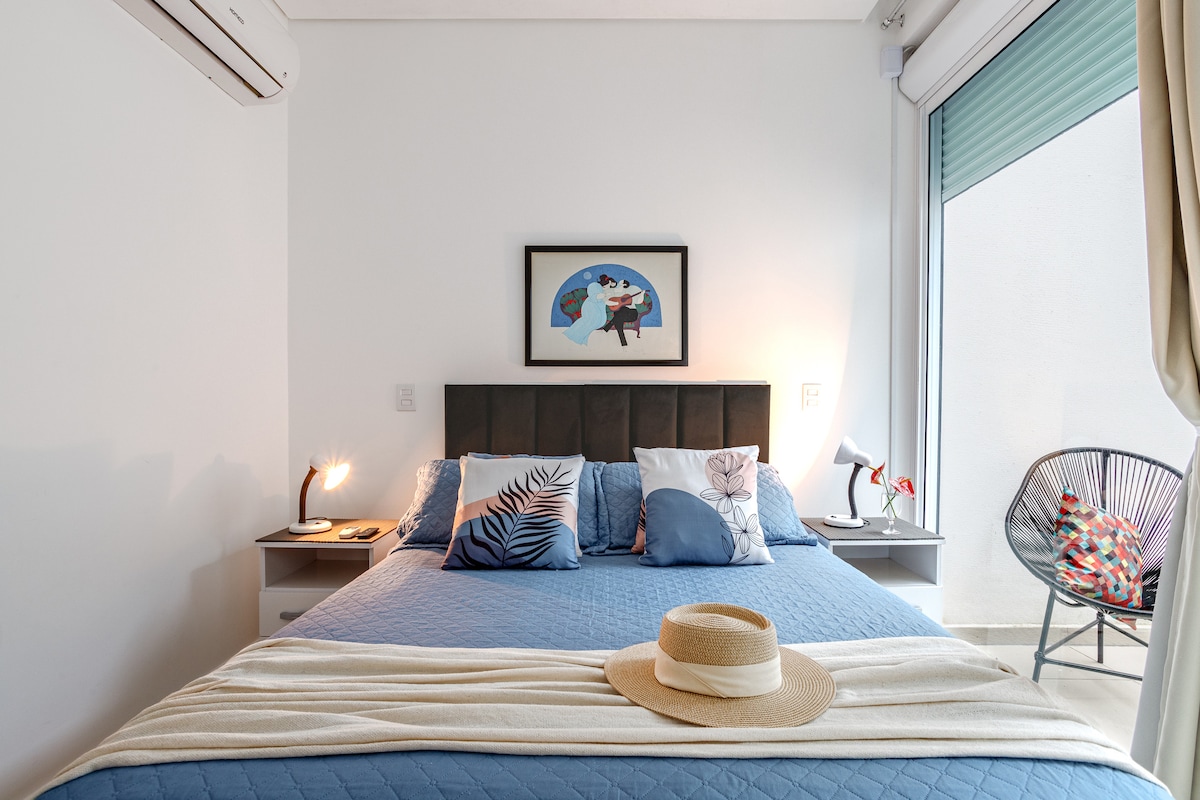 A well-appointed bedroom includes a spacious bed dressed in crisp linens and decorative pillows. A framed artwork is displayed on the wall, and bedside tables hold small lamps. Natural light enters through a glass door that leads to an outdoor space.