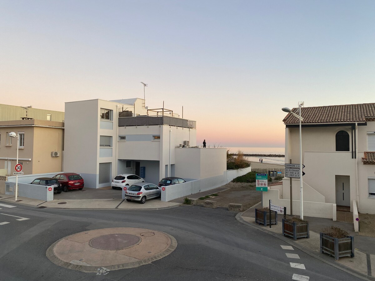 A coastal building is observed from a street view, showcasing a modern architecture with a flat roof. The scene includes vehicles parked near the structure and a glimpse of the beach in the background. The sky transitions from orange to purple as the sun sets.