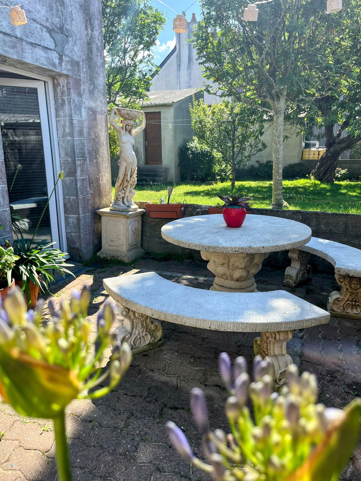 A tranquil outdoor space is presented with a round stone table and curved benches. A potted plant adds a touch of color to the setting. In the background, greenery and a decorative statue create a serene atmosphere, while soft light filters through the trees.