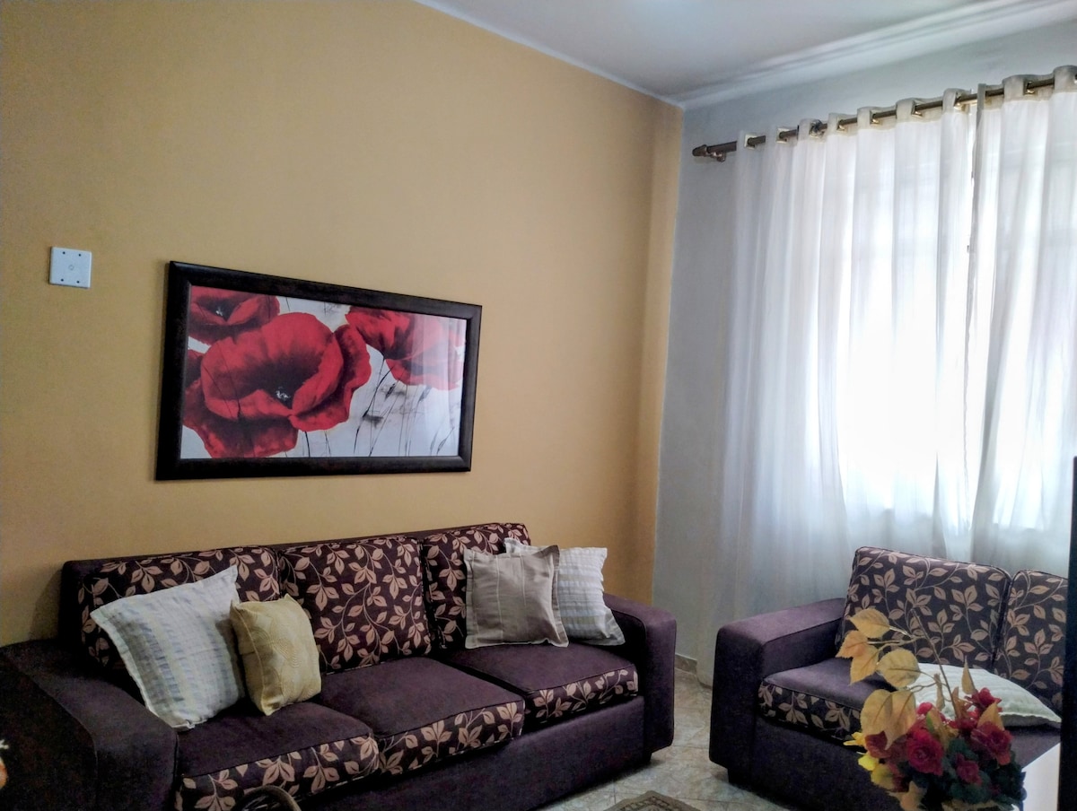 A living area is depicted with two fabric sofas adorned with floral patterns and complemented by soft cushions. A framed artwork featuring red flowers is mounted on an accent wall, while sheer curtains allow natural light to filter through, creating a welcoming ambiance.