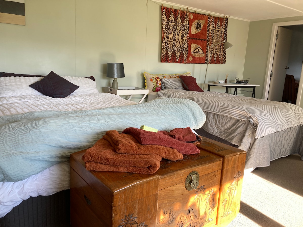 A studio apartment features one queen-sized bed and two single beds arranged to offer flexibility. Cozy blankets and towels are neatly placed on a wooden chest, while a decorative wall hanging adds a touch of character to the room.