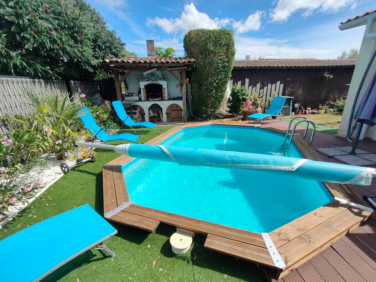 A private area features a rectangular pool surrounded by wooden decking. Lounge chairs in a vibrant blue are positioned nearby. Lush greenery and flowering plants enhance the outdoor ambiance. An outdoor grill can be seen adjacent to the pool, providing a space for gatherings.