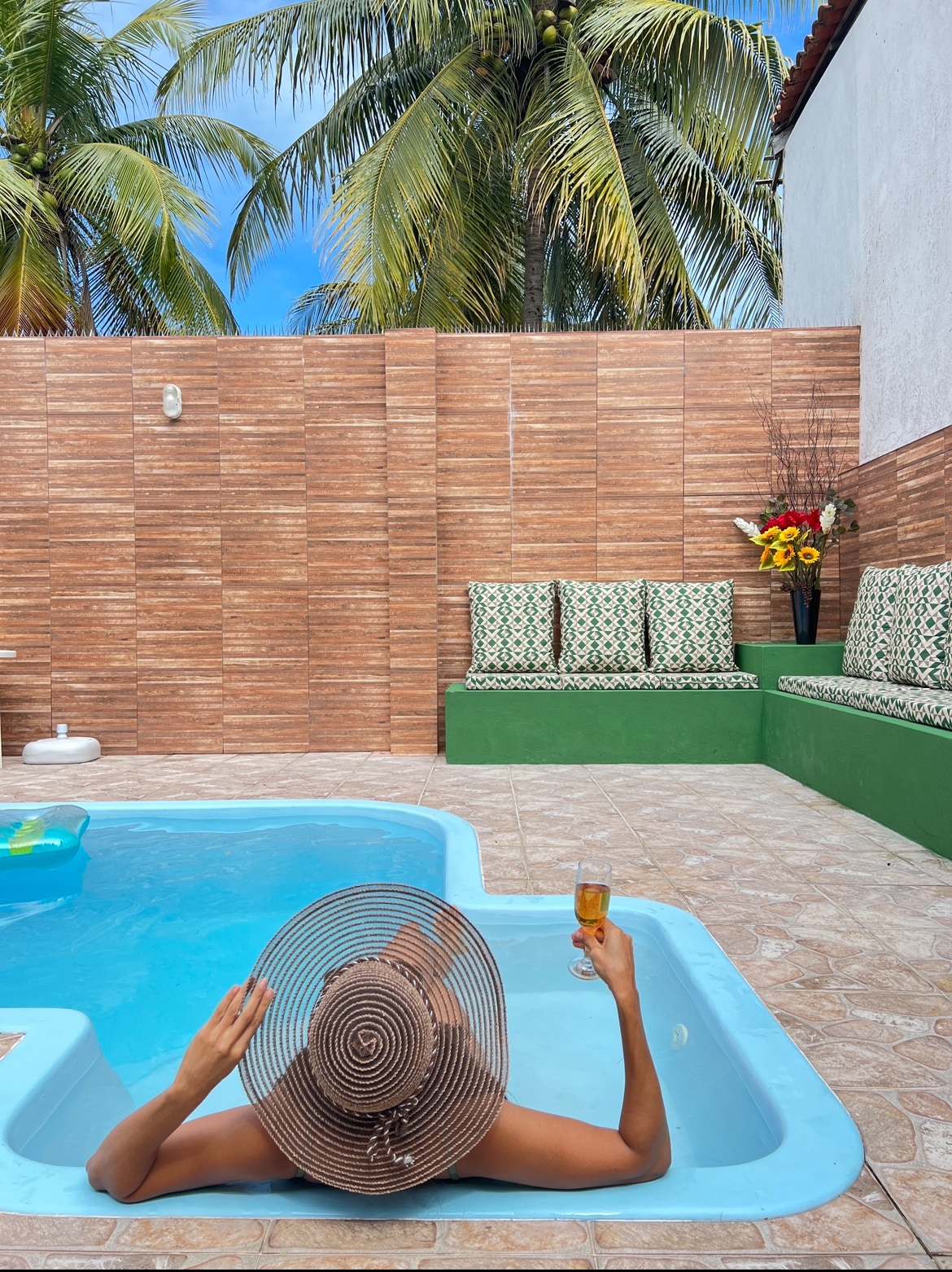 A private swimming pool is showcased, framed by tropical palm trees. Comfortable seating is provided against the wall, with decorative cushions enhancing the area. Natural light illuminates the space, reflecting off the water as a guest relaxes with a beverage.