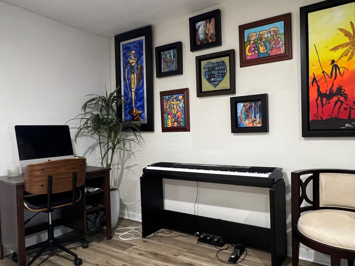A cozy corner of the studio features a digital piano positioned against a decorated wall, adorned with various framed artworks including colorful paintings. A desk with an ergonomic chair sits nearby, accompanied by a potted plant, enhancing the workspace's inviting atmosphere.