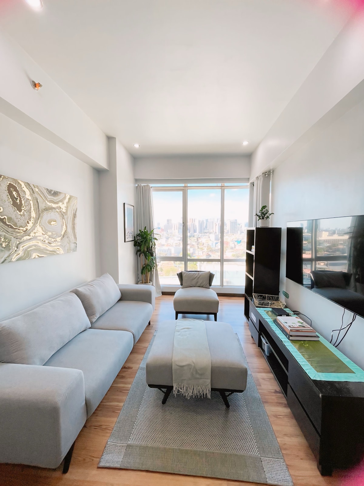 A bright living area features a light grey sofa and an ottoman arranged on a textured rug. A flat-screen TV is mounted on the wall opposite a stylish console, while large windows offer unobstructed views of the cityscape.