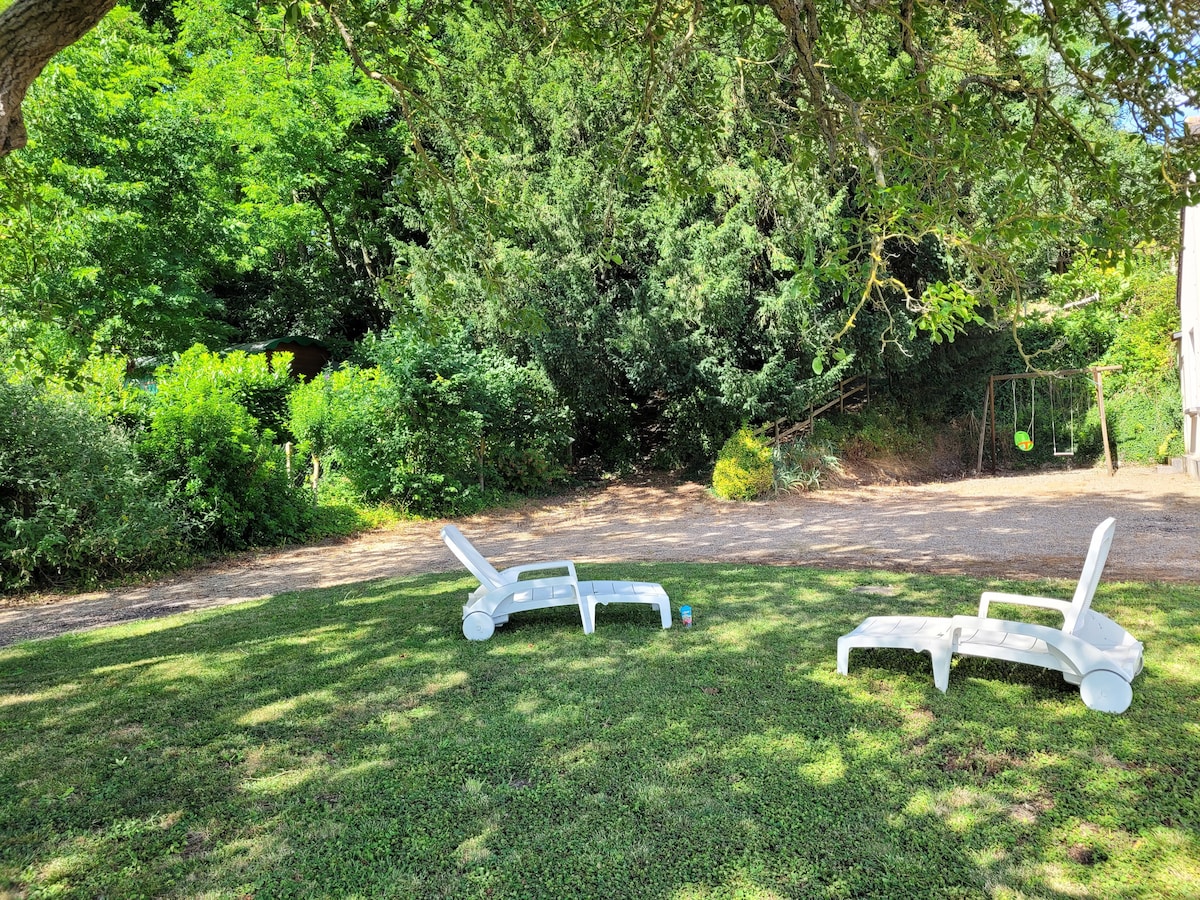 The garden area features two white lounge chairs positioned on lush green grass under the shade of a large tree. In the background, a swing set can be seen, surrounded by dense greenery, creating a serene outdoor space.