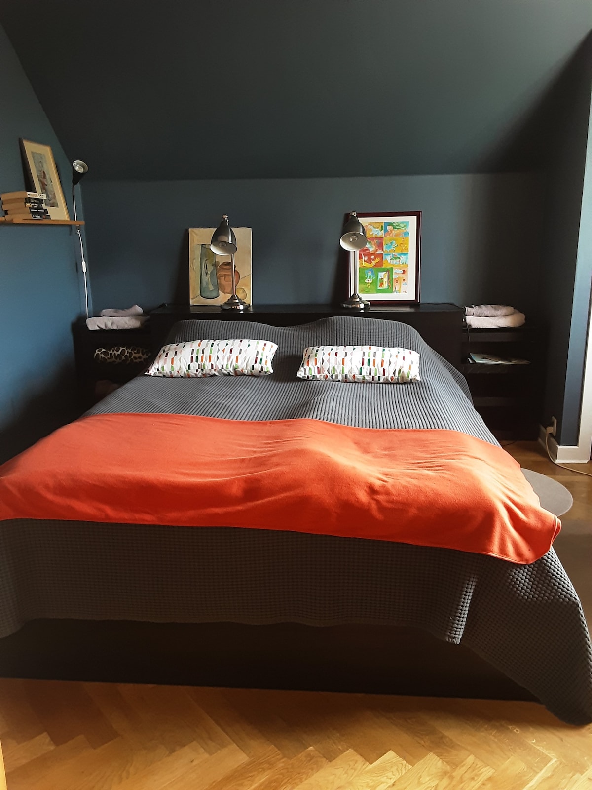 A sleeping area features a spacious bed with patterned pillows. An orange throw blankets the bed, complementing deep blue walls. Two bedside lamps provide illumination, while framed artwork adds color to the space. A small shelf can be seen above the bed.