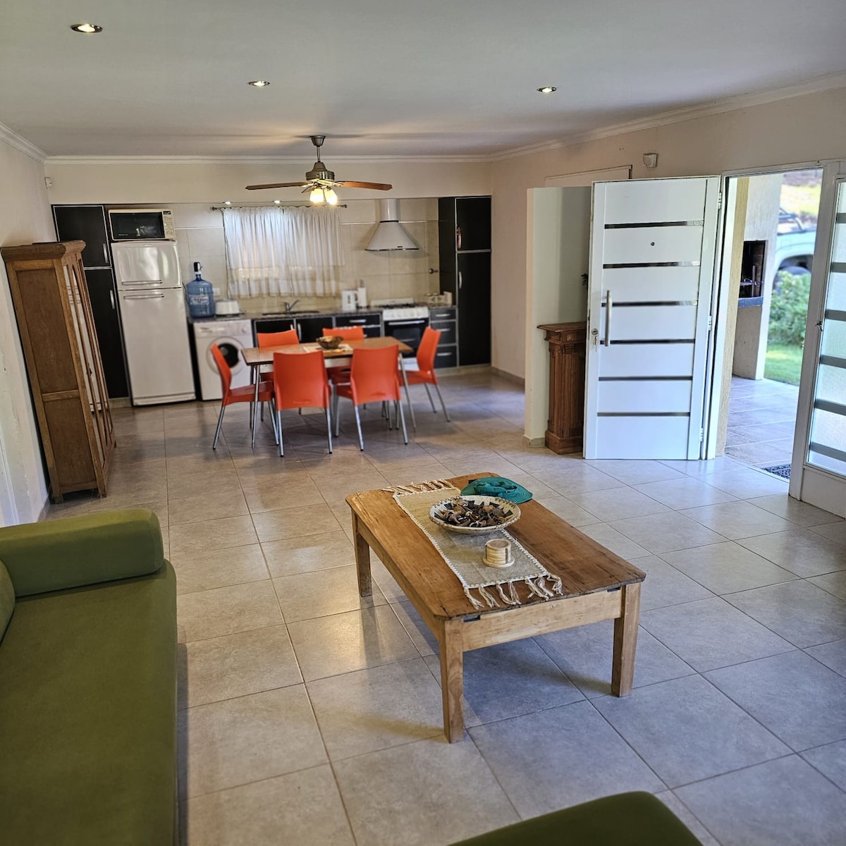 A spacious living area is visible, featuring a green sofa and a wooden coffee table. The open kitchen is equipped with modern appliances, and a dining table with orange chairs is positioned nearby. Bright light enters through glass doors leading to the exterior.