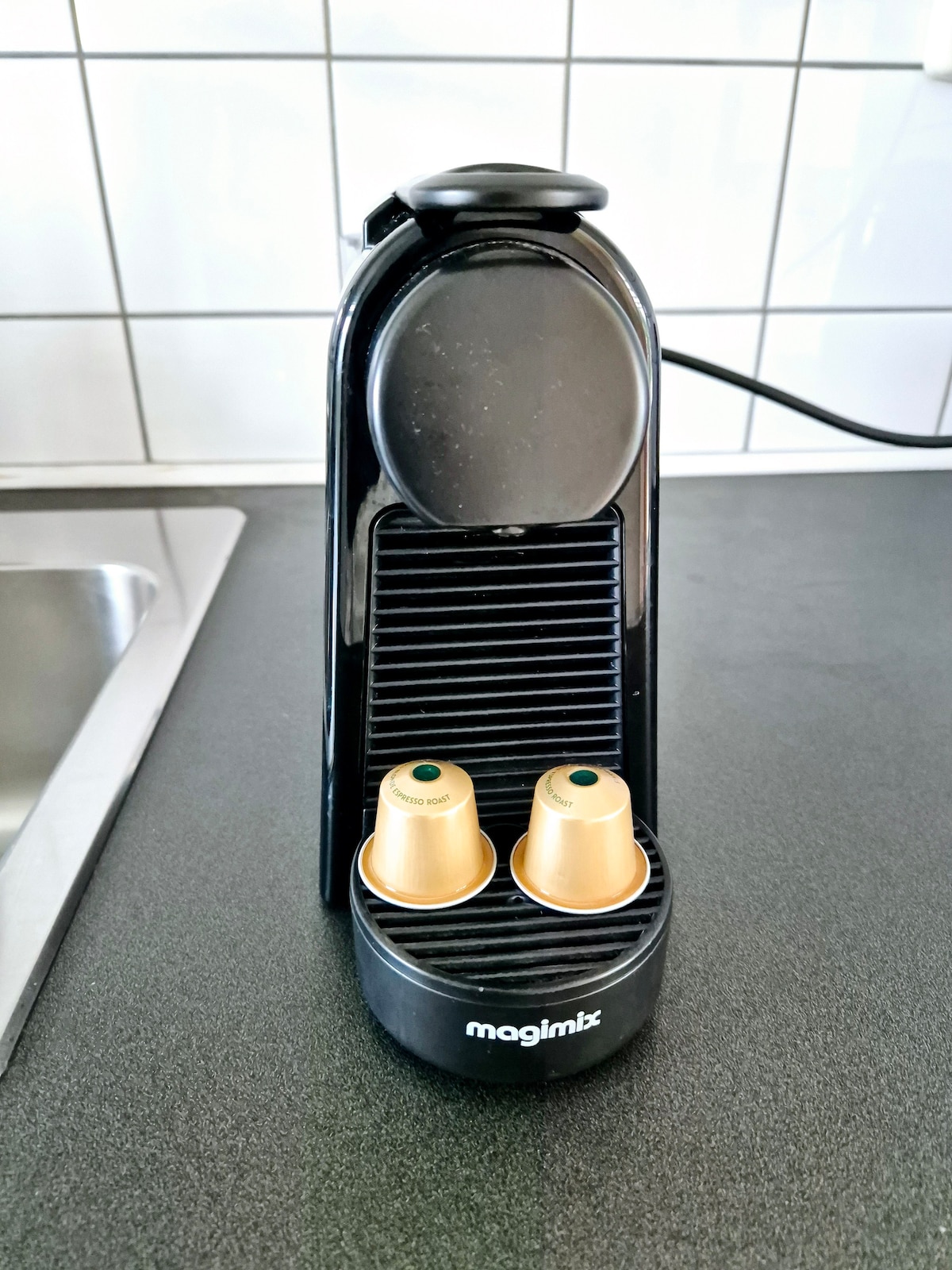 A sleek black coffee machine with a minimalist design sits on a dark countertop. Two golden coffee capsules are positioned in the lower tray, while the machine features a smooth top and a brushed texture on the front panel, highlighting its modern appeal.