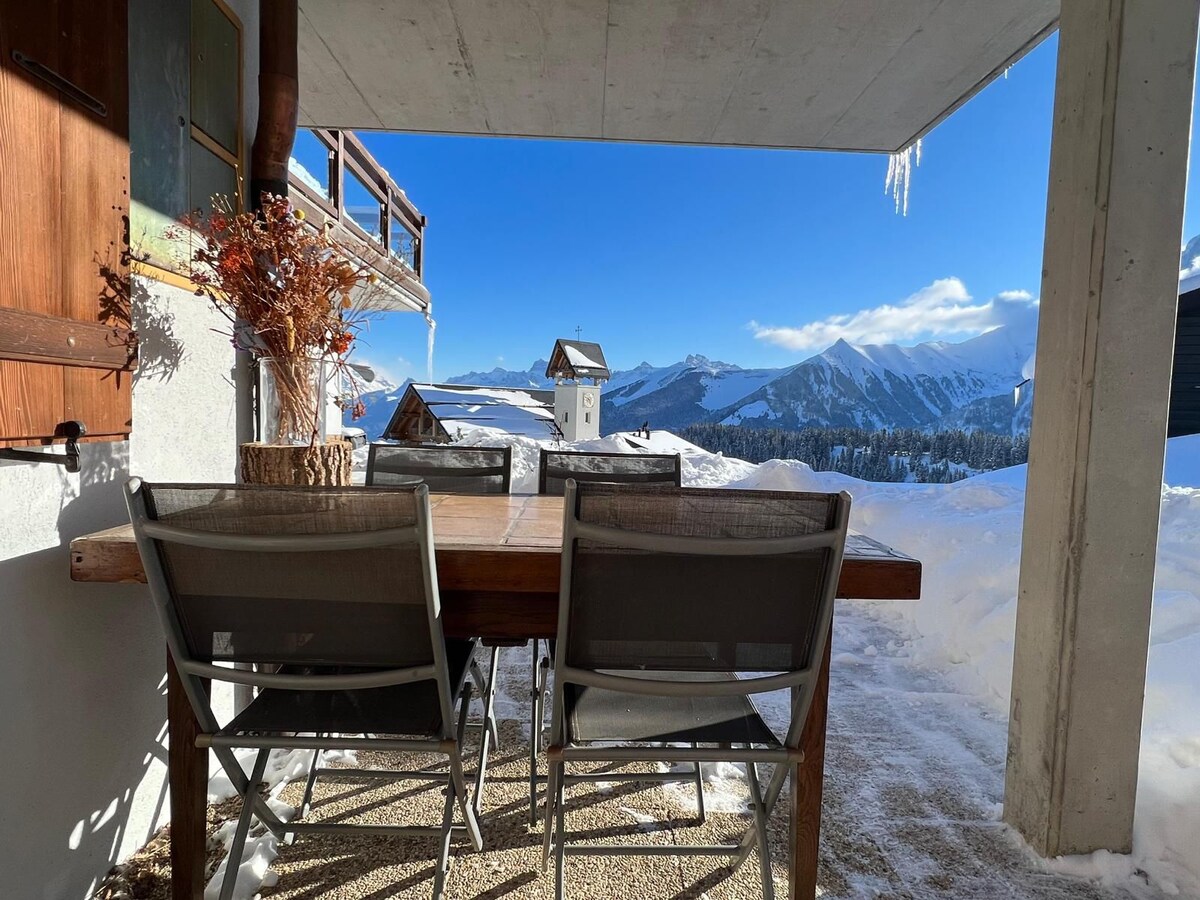 A spacious outdoor terrace is equipped with a large wooden dining table surrounded by several grey chairs. Snow-covered mountains can be seen in the distance, with a clear blue sky above. Icicles hang from the terrace's edge, adding to the winter scene.