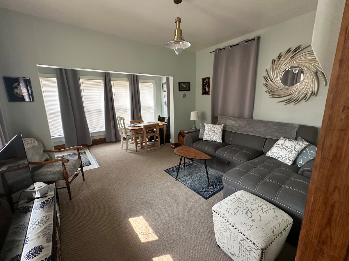 The living room features a cozy sectional sofa and a stylish coffee table, complemented by an inviting dining nook. Natural light fills the space through large windows, while decorative mirrors and curtains add a touch of elegance to the atmosphere.