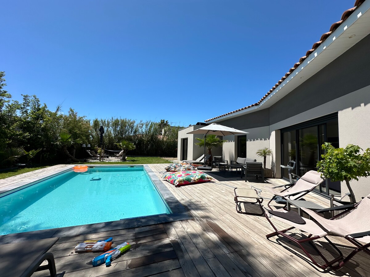 An outdoor terrace features a private swimming pool surrounded by sun loungers and vibrant cushions. Shade is provided by umbrellas. Lush greenery provides a sense of privacy, while a dining area is visible under a covered space adjacent to the pool.