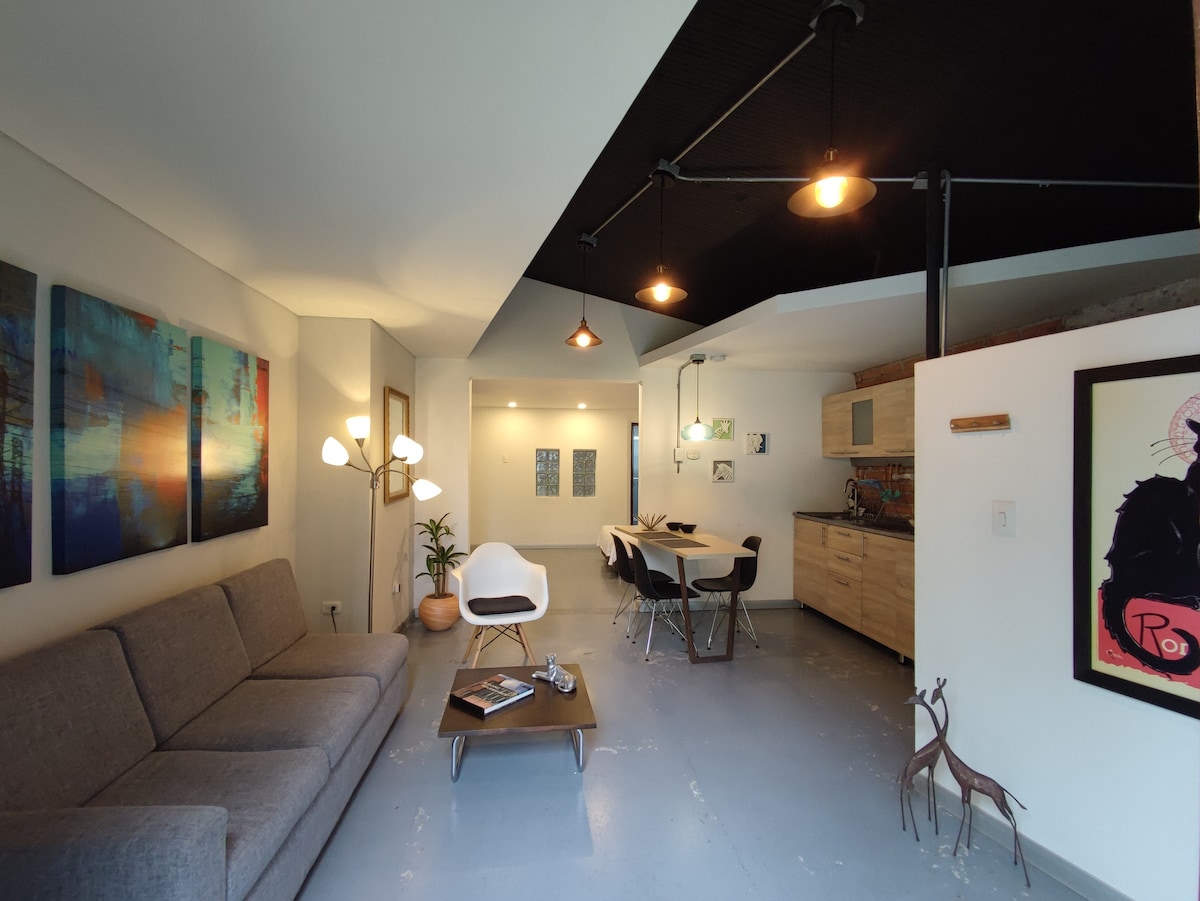A modern living area features a comfortable gray sofa, a stylish white chair, and a coffee table. Artwork adorns the walls, complementing the light fixtures above. An open kitchen area is visible in the background, with dining space furnished for multiple guests.