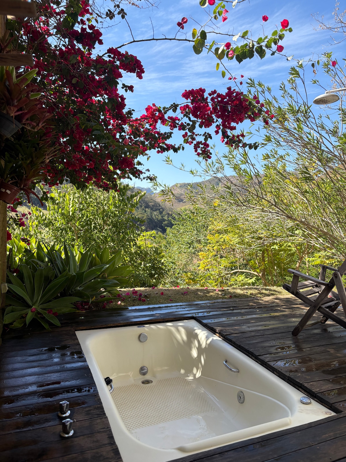 An outdoor hydro bath is set on a wooden deck, surrounded by vibrant green foliage and blooming flowers. The serene natural landscape is visible in the background, enhancing the tranquil atmosphere of the outdoor space.