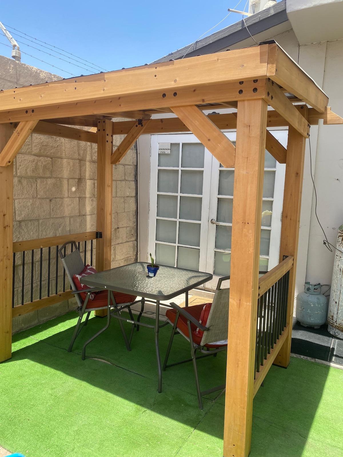 An outdoor seating area features a wooden gazebo structure providing shade. A glass-top table is surrounded by two metal chairs, all positioned on green grass-like flooring. The entrance to the interior is visible through a glass door, leading into the living space.