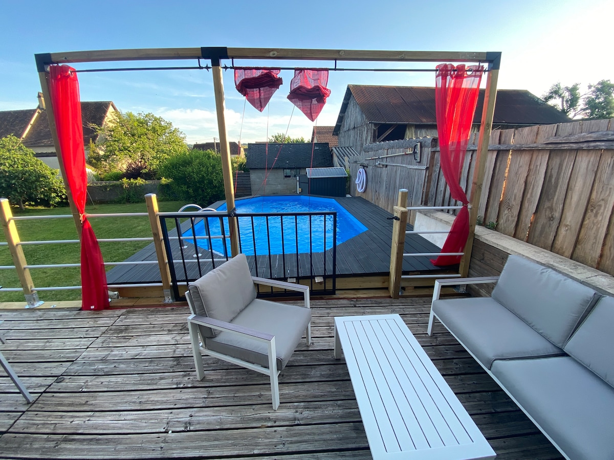 An outdoor deck features two light-colored lounge chairs and a white table, providing a space for relaxation. Behind, a securely fenced pool is visible, framed by vibrant red curtains swaying gently in the breeze. Lush greenery surrounds the area, contributing to an inviting atmosphere.
