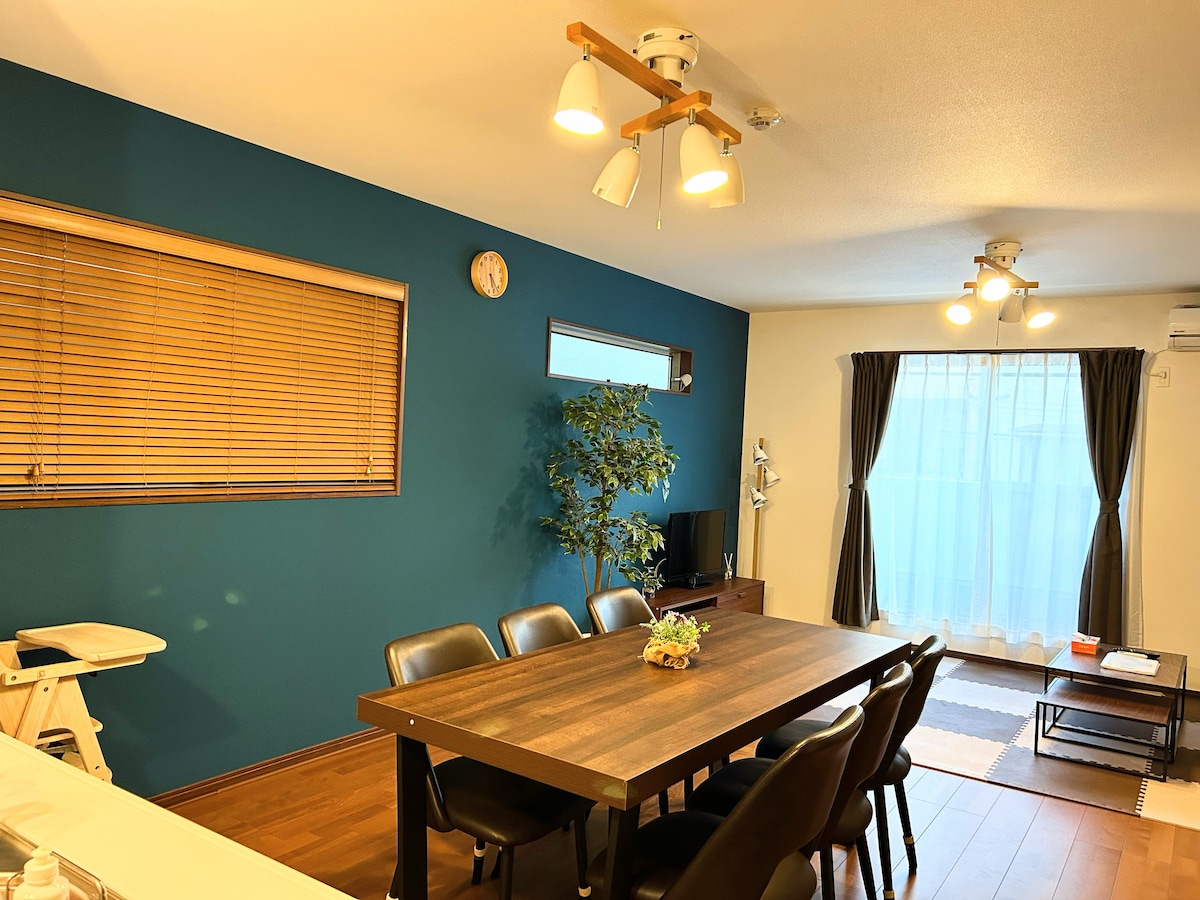 The dining area showcases a wooden table surrounded by black chairs, accommodating multiple guests. A large window covered with light curtains allows natural light to fill the space. A green plant and wall clock add functional touches, against a contrasting blue wall.