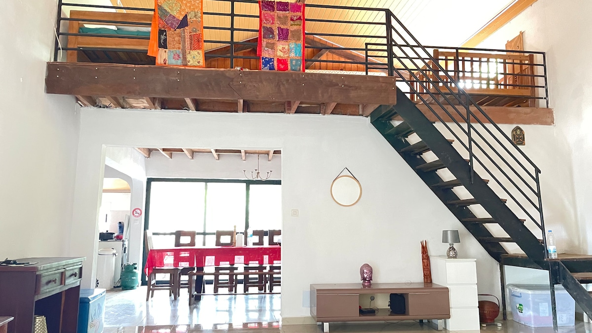 A spacious interior features a staircase leading to a loft area with vibrant wall hangings. The ground level includes a dining space with a long table and chairs, alongside a contemporary low television stand. Large windows allow natural light to fill the room.