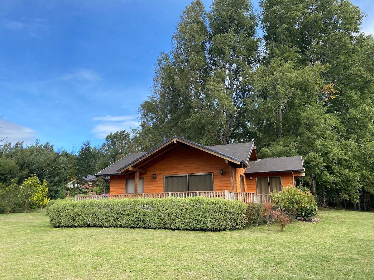 A wooden house is framed by a well-maintained lawn and native trees, providing a natural setting. The exterior features large windows that allow ample light, and decorative shrubs enhance the landscape. The presence of greenery creates a peaceful and inviting environment.