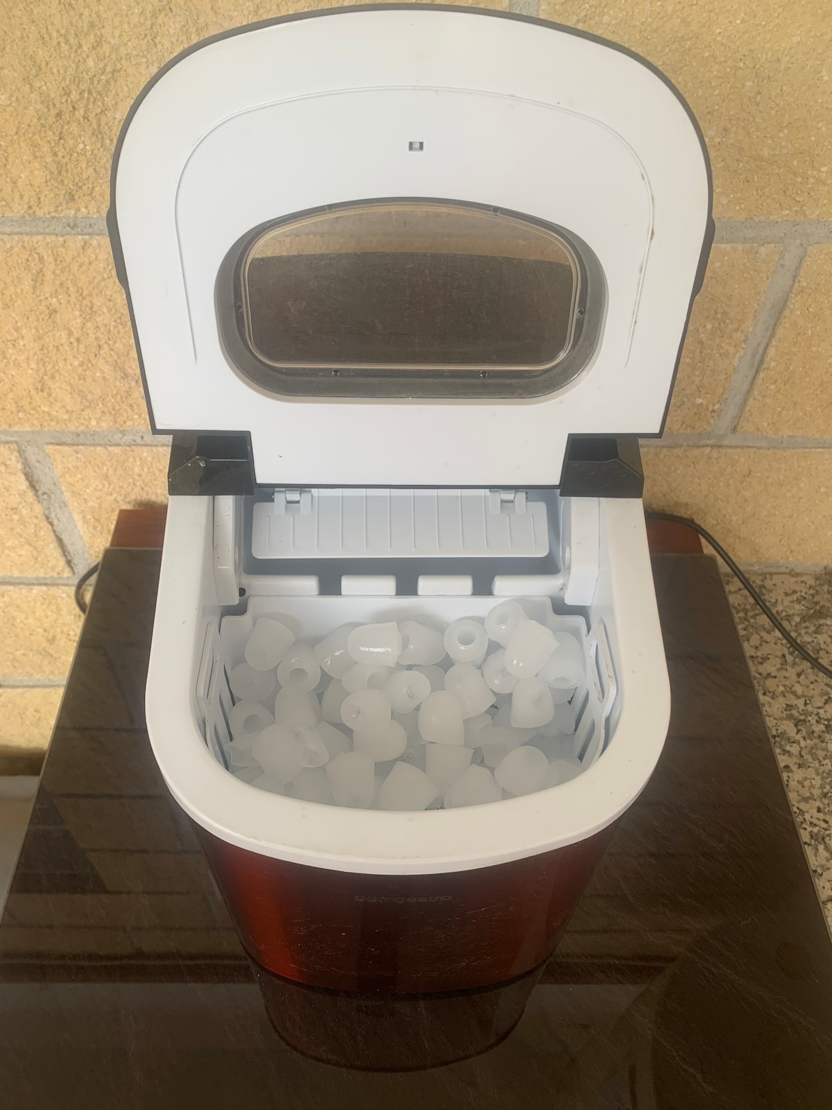 An ice maker is shown with its lid open, displaying a full compartment of ice cubes. The machine features a clear viewing window on the lid, allowing for visual access to the ice inside. It rests on a dark countertop, surrounded by a light-colored wall.