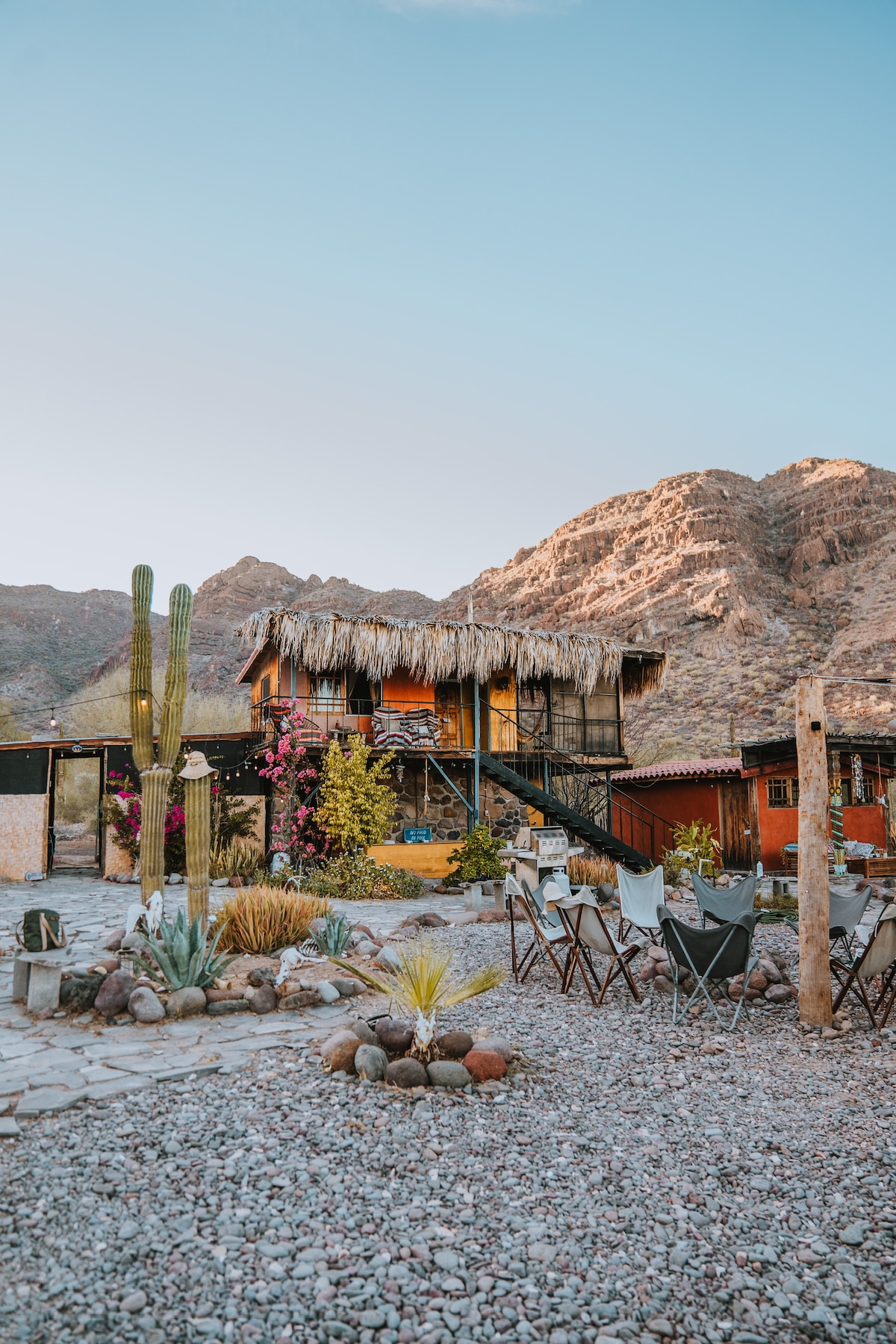 A rustic glamping site is nestled against a backdrop of mountains, showcasing a multi-level structure with a thatched roof. Surrounding flora includes cacti and colorful flowers, while a pebbled path leads to a cozy seating area with chairs, inviting relaxation in nature.
