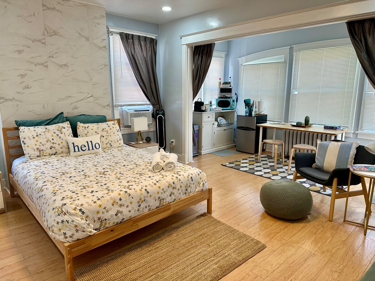 A bright and airy guest suite features a spacious bed adorned with patterned bedding. A cozy reading area with a small table is visible, along with a kitchen space in the background. Natural light streams through large windows, enhancing the warm ambiance.