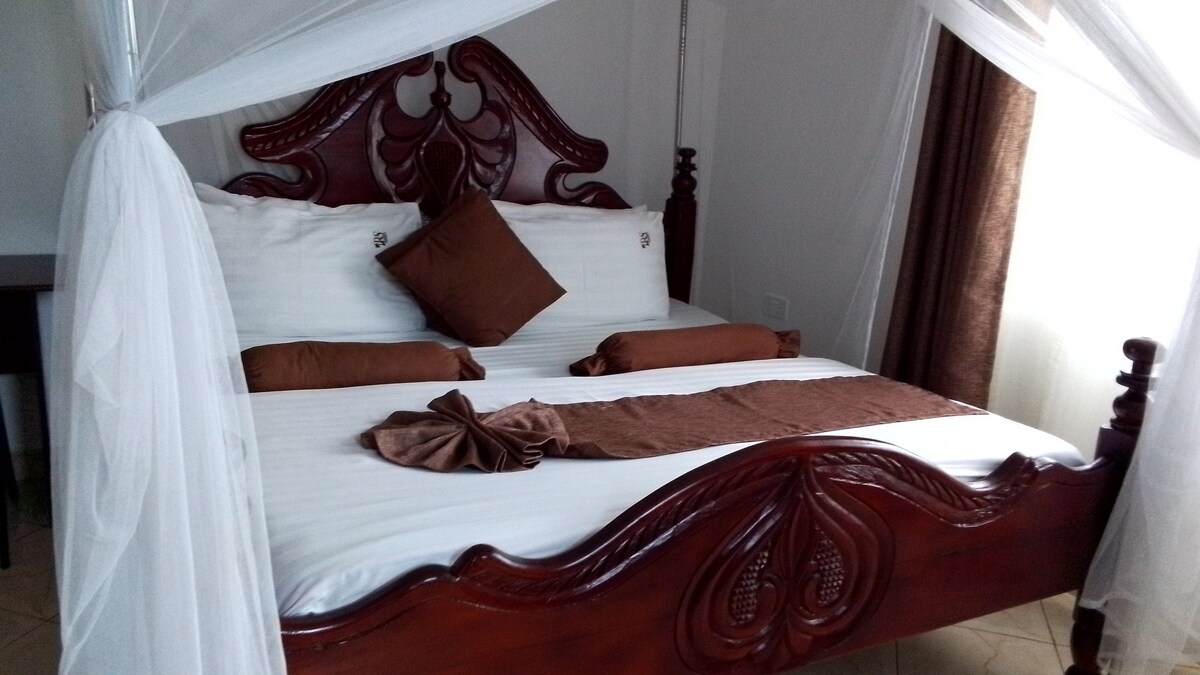 A wooden bed with an intricately carved headboard is complemented by a soft white canopy. The bed is dressed in white linens and includes decorative brown pillows and a coordinated throw, creating a comfortable sleeping space. Natural light filters through nearby curtains.