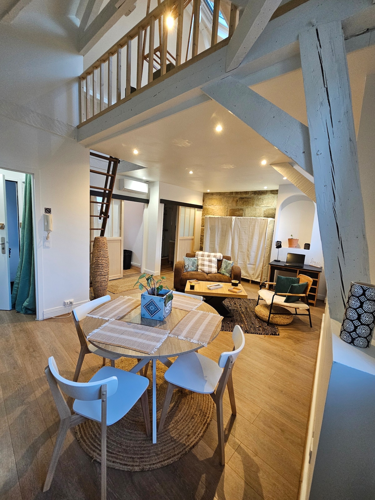 A bright and open living space features a dining table set for four, surrounded by modern chairs. A cozy seating area includes a patterned sofa and a small coffee table, while light filters through the windows, highlighting the wooden beams above.