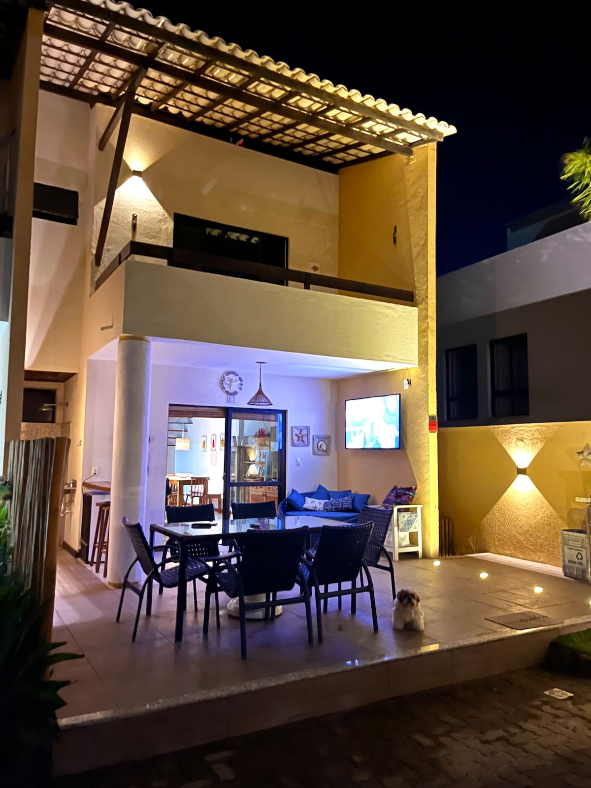 A duplex structure is illuminated at night, showcasing a spacious outdoor patio furnished with black dining chairs and a small table. A cozy seating area is visible, with decorative accents and warm lighting enhancing the ambiance. A small dog is seen on the patio floor.
