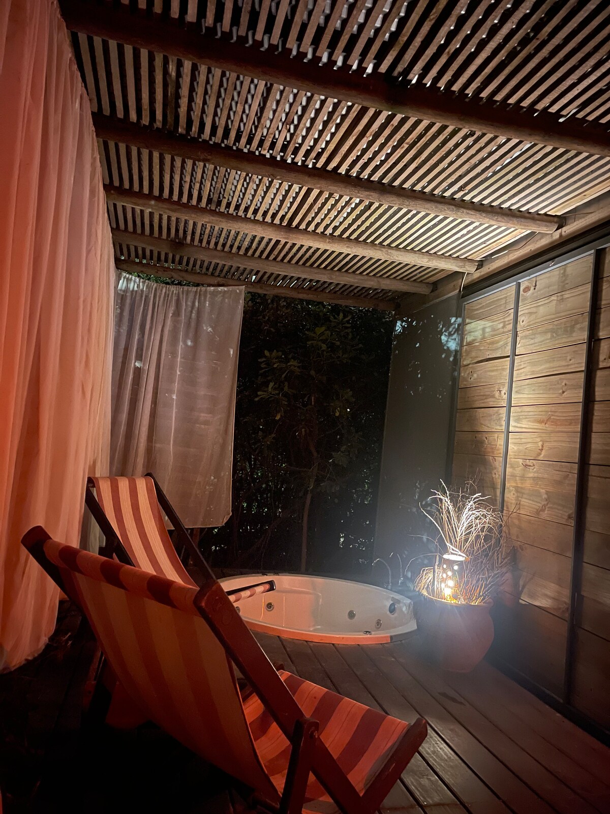 An outdoor space features two striped deck chairs positioned beside a private jacuzzi. Soft light emanates from a nearby lantern, illuminating the area. Sheer curtains provide a touch of privacy, while shadowy greenery enhances the serene atmosphere.