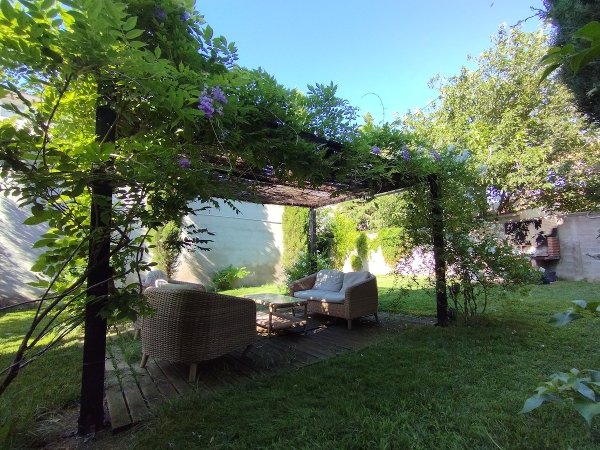A shaded seating area is positioned under a pergola adorned with lush foliage. Two comfortable chairs surround a small table, creating a serene spot for relaxation. The green lawn adds to the inviting outdoor ambiance, with surrounding plants contributing to the tranquil setting.