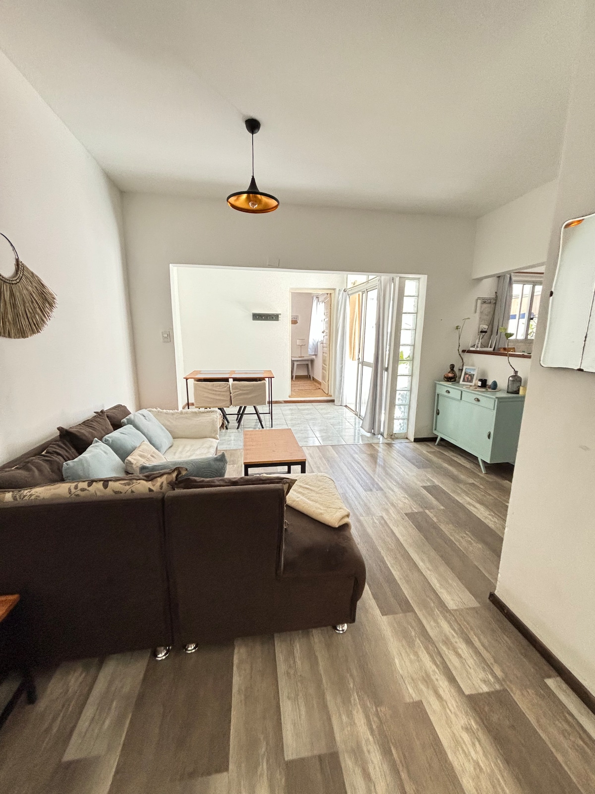 A comfortable living area features a dark-colored sofa with decorative cushions, positioned beside a wooden coffee table. Light hardwood flooring extends throughout the space, illuminated by natural light from open doors leading to an outdoor area. A small, light-colored dining table can be seen in the background.