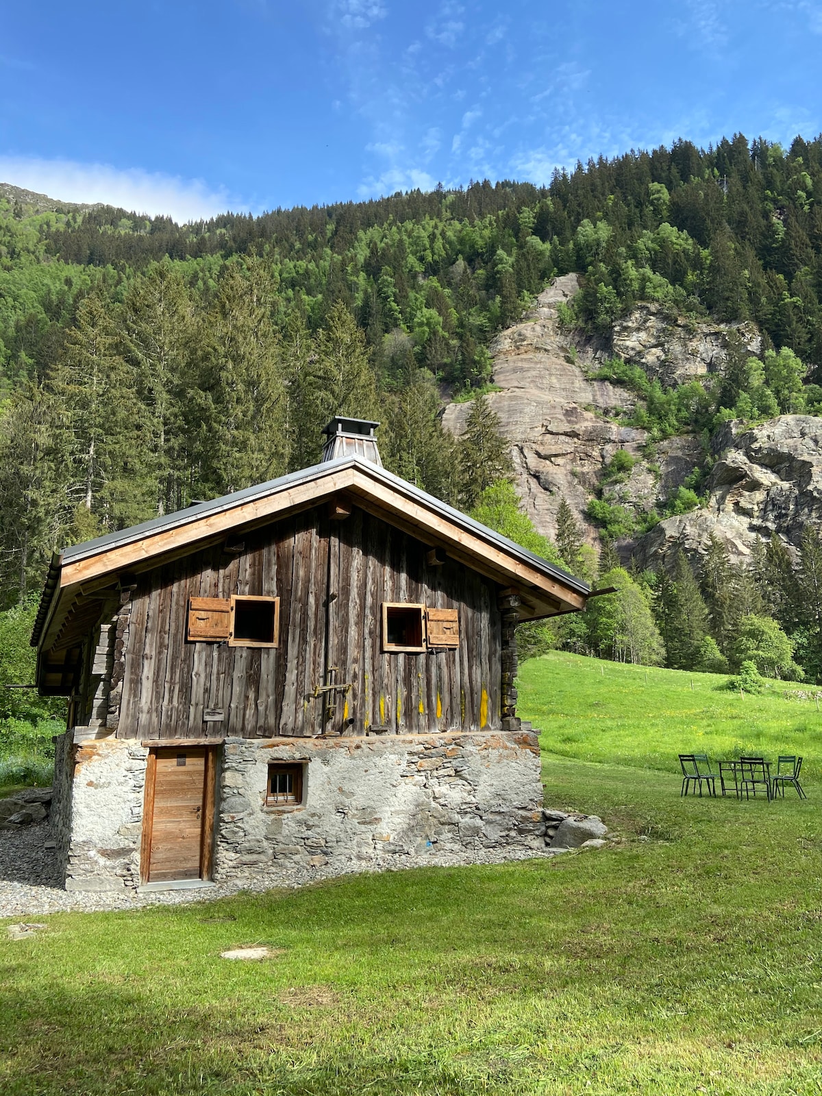 The rustic cabin features a wooden exterior with stone accents, nestled in a lush green landscape. Tall trees and rocky cliffs provide a natural backdrop, while a small outdoor seating area is visible nearby, inviting moments of relaxation in the serene surroundings.