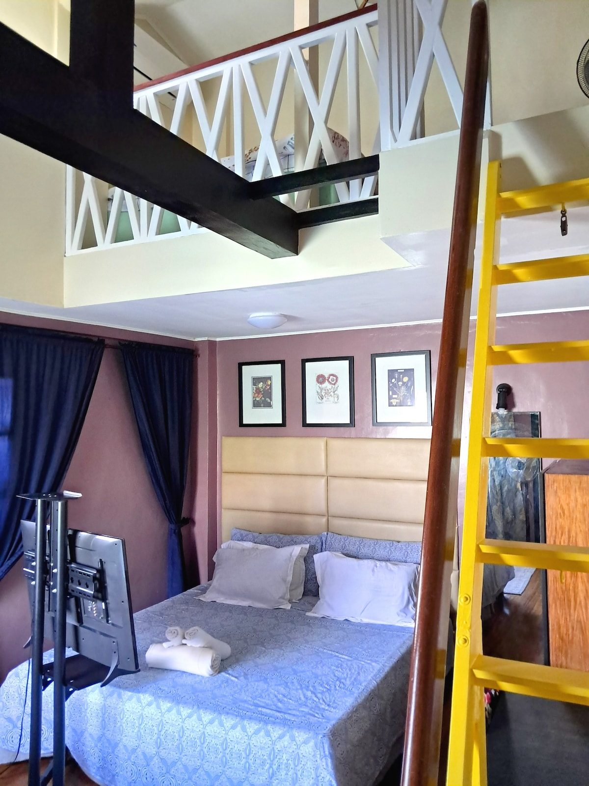 A spacious sleeping area features a comfortable bed with fresh linens. Above, a loft area is visible. A flat-screen TV stands on a stand nearby, and framed artwork accentuates the walls. Soft lighting complements the elegant color scheme, while curtains flank the window.