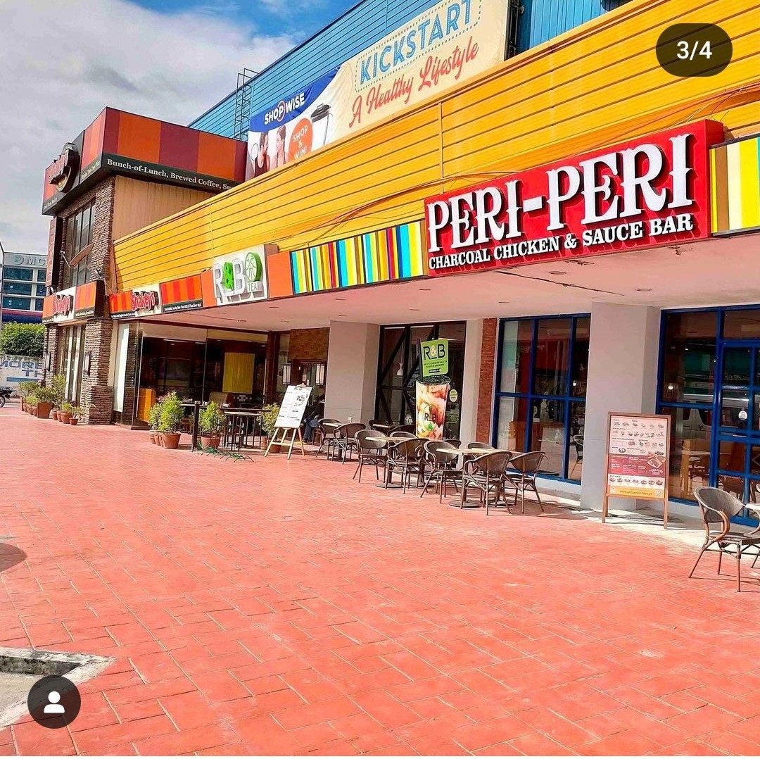 A vibrant outdoor area features a series of colorfully accented storefronts, including a charcoal chicken restaurant. Several tables and chairs are arranged outside, inviting guests to dine al fresco. The setting is brightened by natural light under a clear sky.