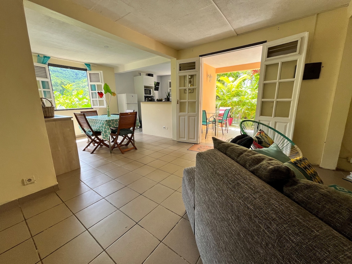 An open living area is displayed, featuring a comfortable gray sofa and a patterned dining table. Bright natural light streams in through large glass doors, leading to an outdoor terrace with colorful chairs. A view of lush greenery is visible beyond the entrance.
