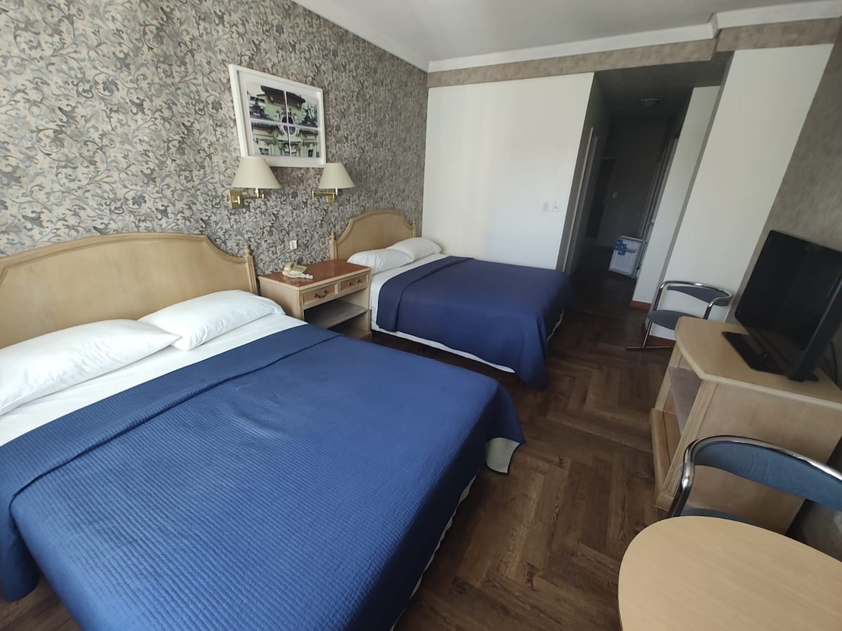 The room features two spacious beds with blue bedding. A floral accent wall adds a touch of character. A wooden nightstand and a small desk with a chair are present. A TV is positioned on a wooden unit, with subtle lighting enhancing the overall ambiance.