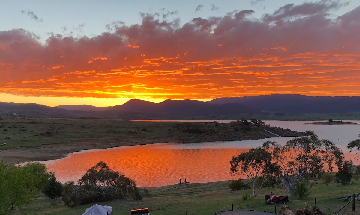 A vibrant sunset casts hues of orange and pink across the lake, with silhouettes of distant mountains framing the scene. Gentle waves reflect the colors, while trees line the shore, providing a natural backdrop to the serene lakeside environment.