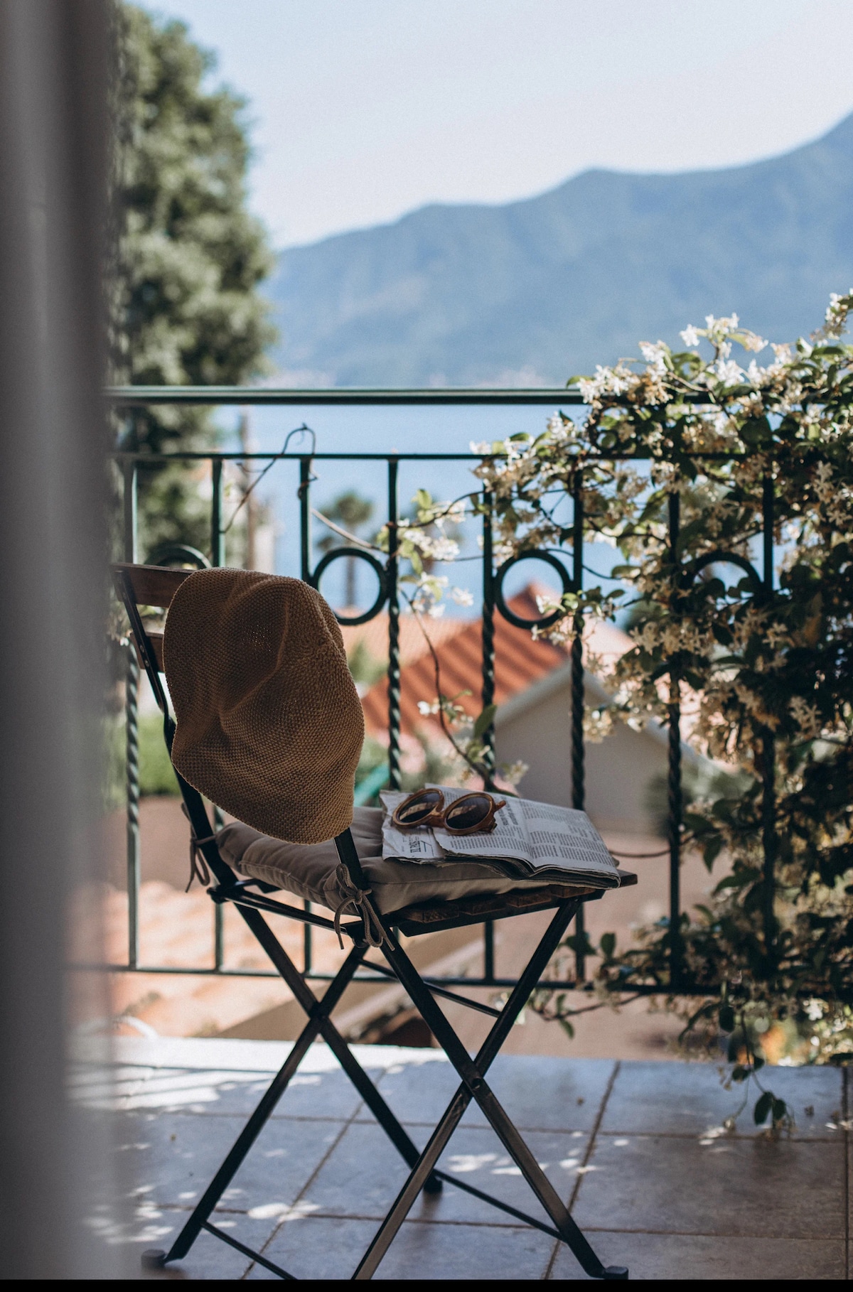 A collapsible chair is positioned on a terrace with a natural view. A sunhat rests on the chair along with a pair of sunglasses and a magazine. The surroundings include lush plants and the distant outline of mountains, enhancing the serene setting of the outdoor space.