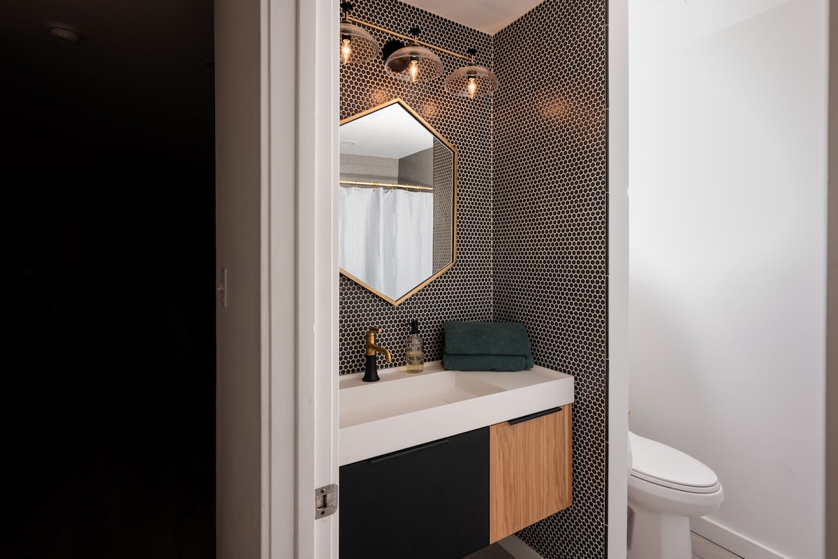 The bathroom features a modern design with a hexagon mirror above a sleek vanity. A stylish blend of black and wood cabinetry is complemented by a decorative towel. The space is illuminated by pendant lighting against a contrasting tile wall.