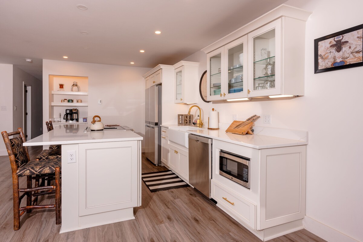The kitchen features modern cabinetry with glass-front shelves showcasing dishware. Stainless steel appliances, including a refrigerator and microwave, are integrated seamlessly. A spacious island offers seating, and a neutral color palette complements the sleek countertops.